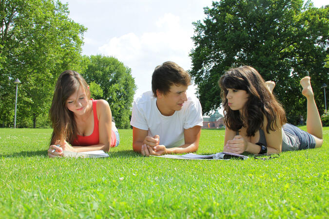 Zwei Mädchen und ein Bursch liegen im Park auf der Wiese und lernen