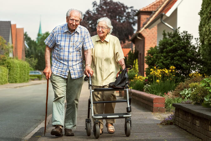 Seniorenpaar mit Stock und Rollator gehen durch eine Wohnsiedlung