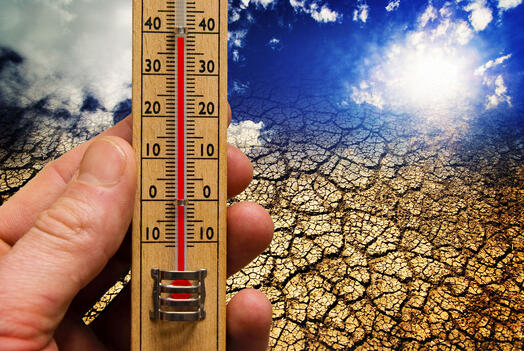 Symbolbild für Klimawandel und globale Erwärmung: Jemand hält ein Thermometer in der Hand, das 39° anzeigt; im Hintergrund ausgedörrte Erde und ein Himmel mit Sonne und Wolken